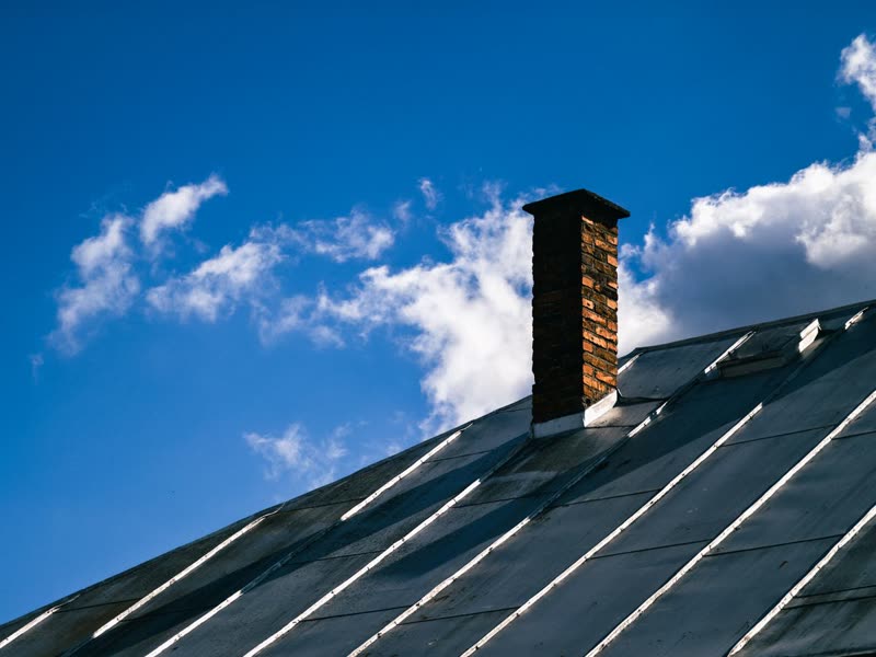 Dark standing seam metal roof with a brick chimney under a blue sky
